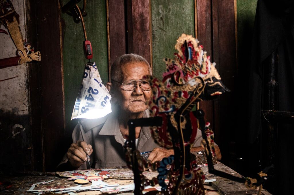 An elderly man crafts intricate wayang puppets in a traditional Indonesian setting.