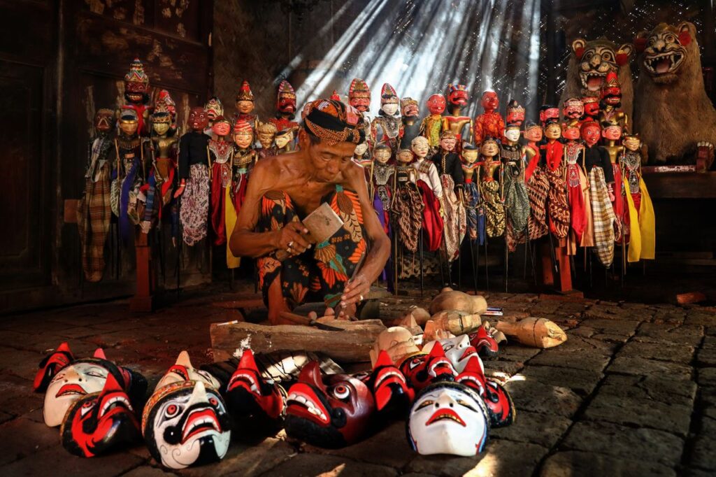 A craftsman in Indramayu, Indonesia, skillfully creates traditional masks amidst wayang puppets.