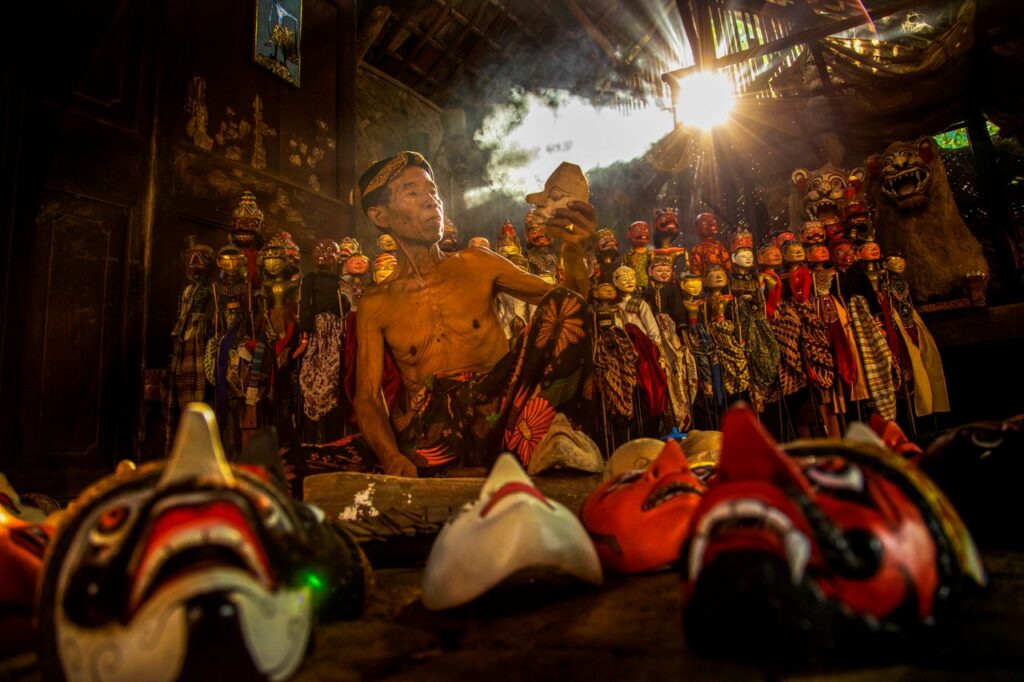 A skilled artisan crafting masks in a traditional workshop in West Java, Indonesia, surrounded by vibrant masks.