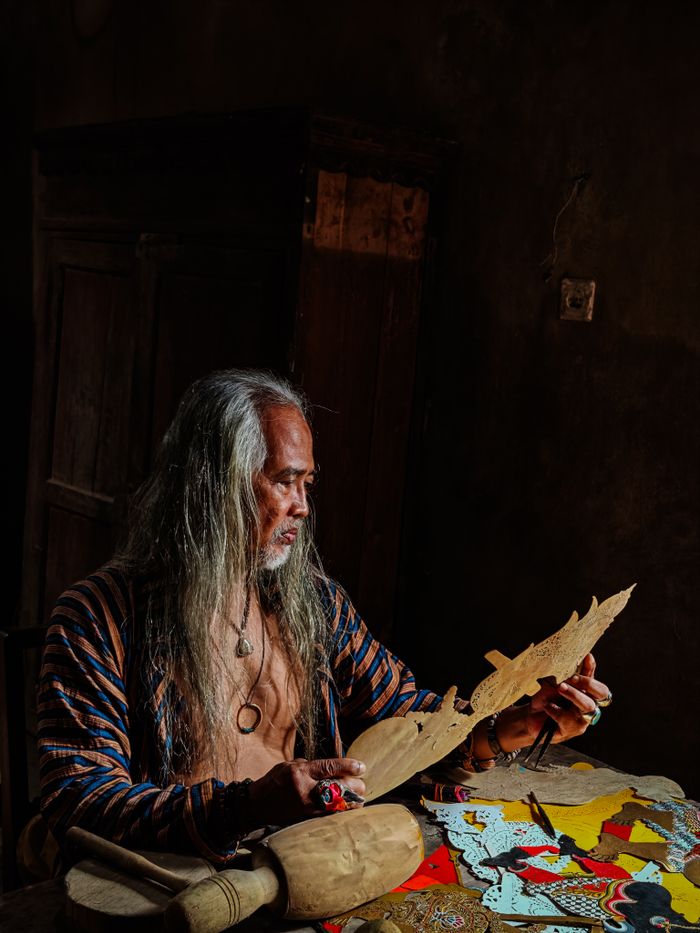 An artisan skillfully crafts shadow puppets at a wooden table in a dimly lit room, showcasing traditional craftsmanship.